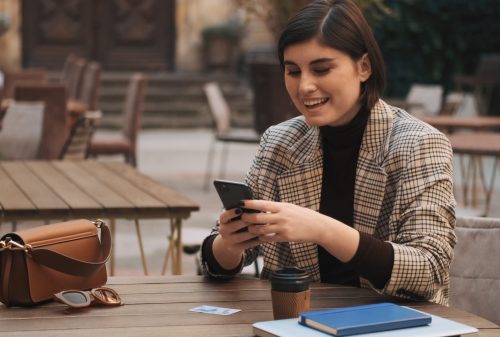 flexible-booking Attractive cheerful female entrepreneur looking happy texting on smartphone during coffee break in courtyard of cafe